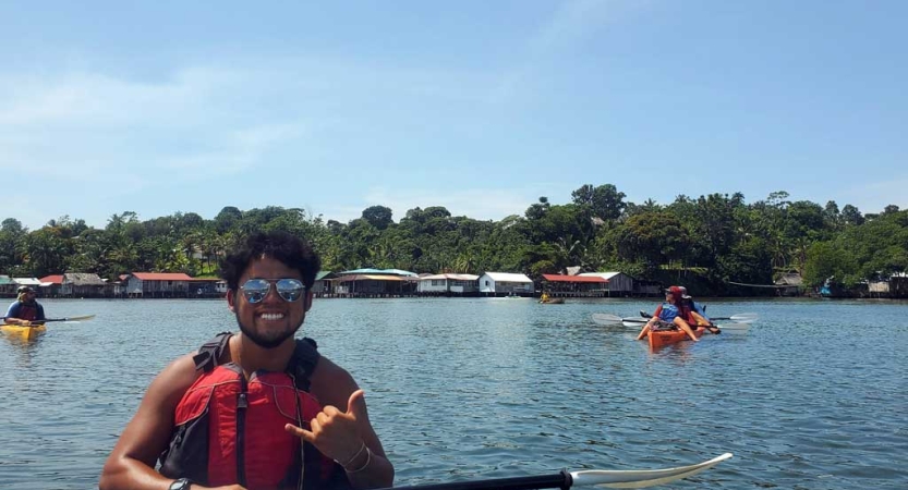 A person wearing a lifejacket sits in a kayak, sits in a kayak, smiles and gives the "hang loose" sign